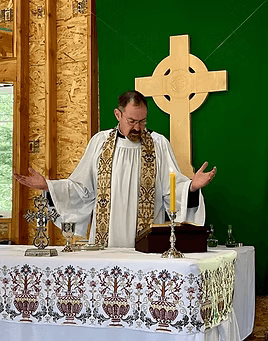 Man wearing liturgical vestments standing behind a church altar with hands raised in prayer, with a chalice, candle, and communion vessels on the altar and a large cross behind him.